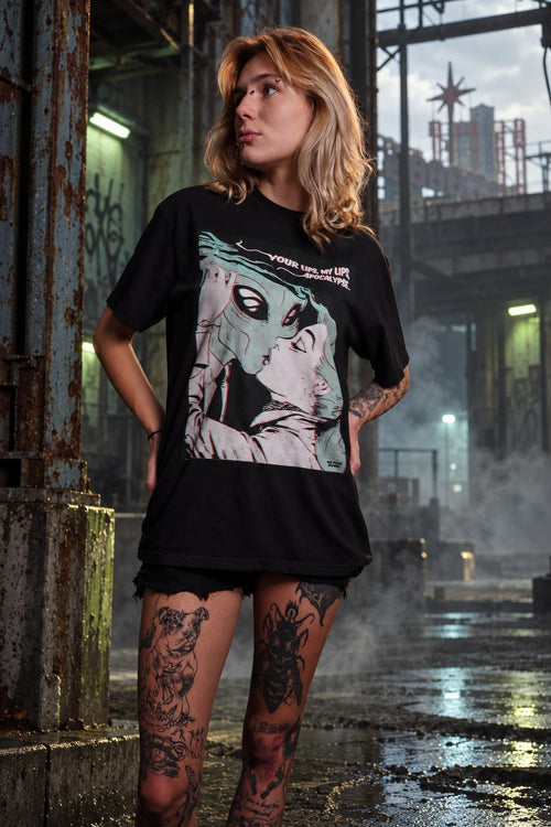 Model wearing the OLD VEGAS Alien Kiss black graphic tee, showing the alien and human kiss artwork in an industrial setting.